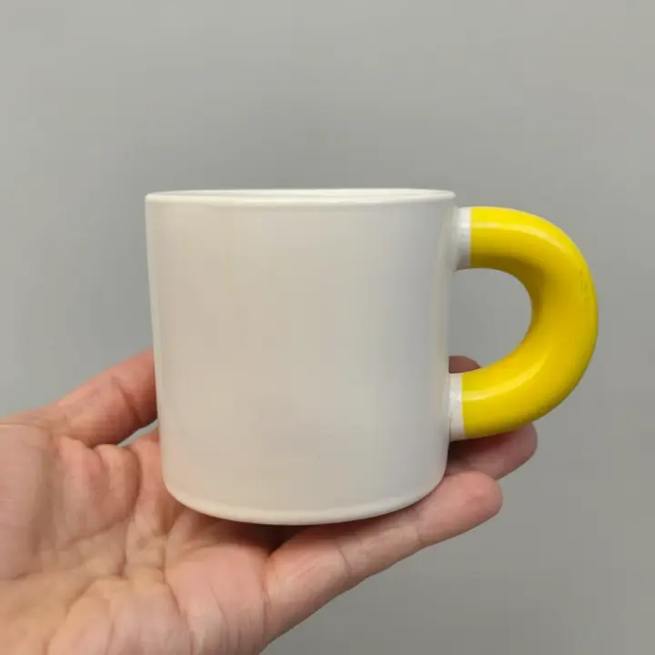 An elegant ceramic mug being held by a hand from below, showing its medium size. The mug has a simple, artistic handcrafted design that combines simplicity and elegance. The background is a neutral light gray that hig.webp