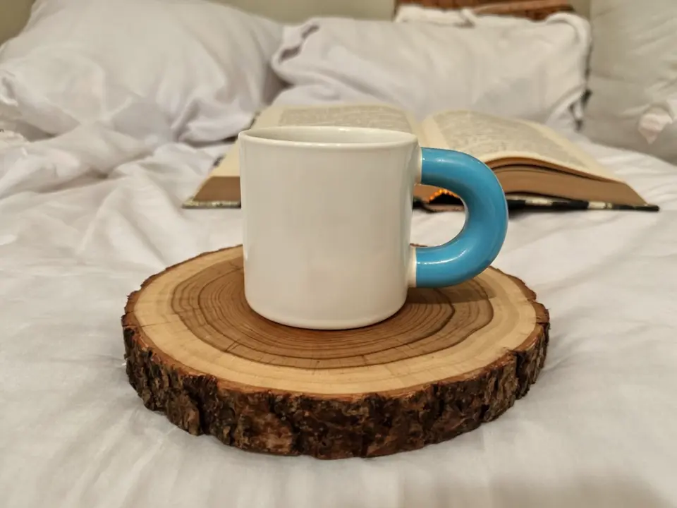 A white ceramic mug with a bright blue handle placed on a circular wooden coaster cut from a tree trunk showing growth rings. The background consists of soft white fabric like a bed or couch, with an open book visible.webp