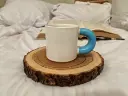 A white ceramic mug with a bright blue handle placed on a circular wooden coaster cut from a tree trunk showing growth rings. The background consists of soft white fabric like a bed or couch, with an open book visible.webp